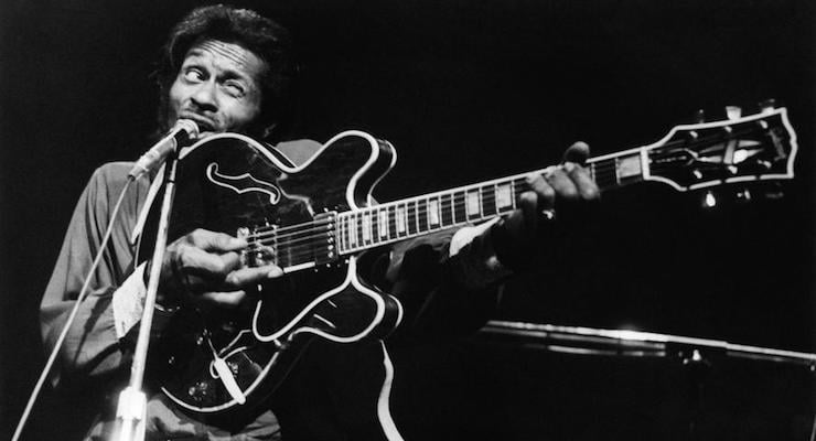 Rock 'n' roll legend Chuck Berry in a black and white promo photo holding his guitar and winking.