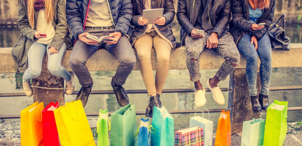 Group of friends sitting outdoors with shopping bags; several people holding smartphones and tablets. (Photo: AdobeStock/ OneInchPunch/PPD)