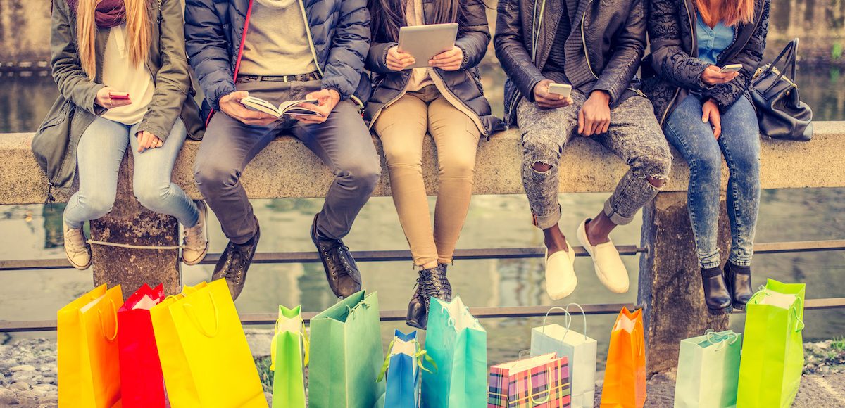 Group of friends sitting outdoors with shopping bags; several people holding smartphones and tablets. (Photo: AdobeStock/ OneInchPunch/PPD)