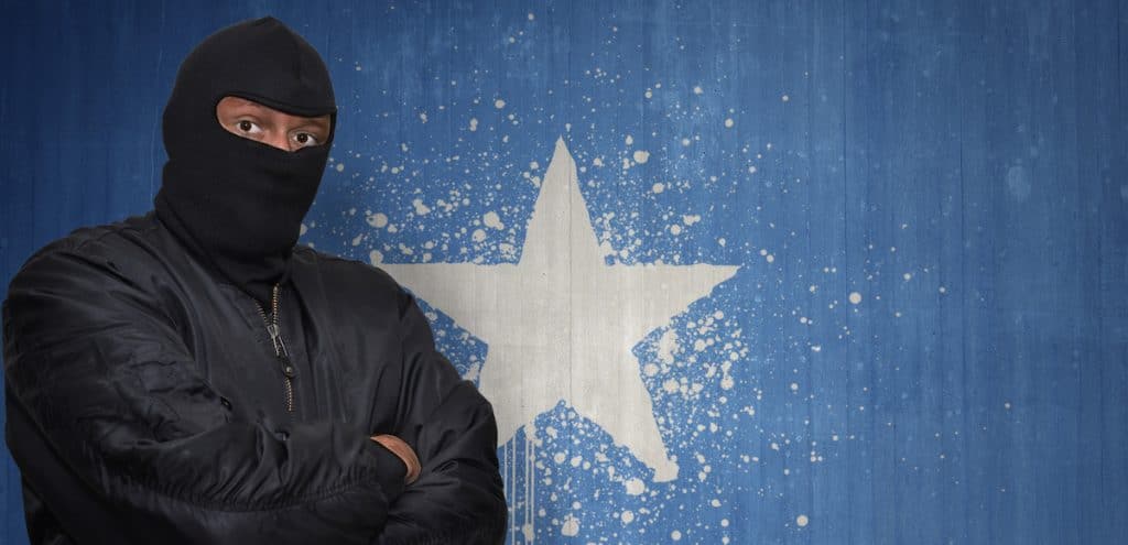 A man posing as an Islamic terrorist in a mask standing in front of a wall painted with the national flag of Somalia. (Photo: AdobeStock)