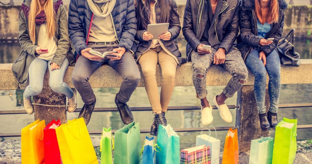 Group of friends sitting outdoors with shopping bags; several people holding smartphones and tablets. (Photo: AdobeStock/ OneInchPunch/PPD)