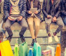Group of friends sitting outdoors with shopping bags; several people holding smartphones and tablets. (Photo: AdobeStock/ OneInchPunch/PPD)