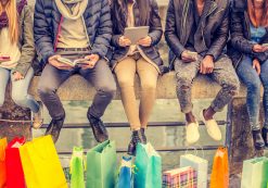 Group of friends sitting outdoors with shopping bags; several people holding smartphones and tablets. (Photo: AdobeStock/ OneInchPunch/PPD)