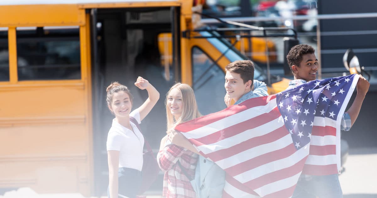 A group of American public school teenage students with an American flag in front of school bus. (Photo: AdobeStock)