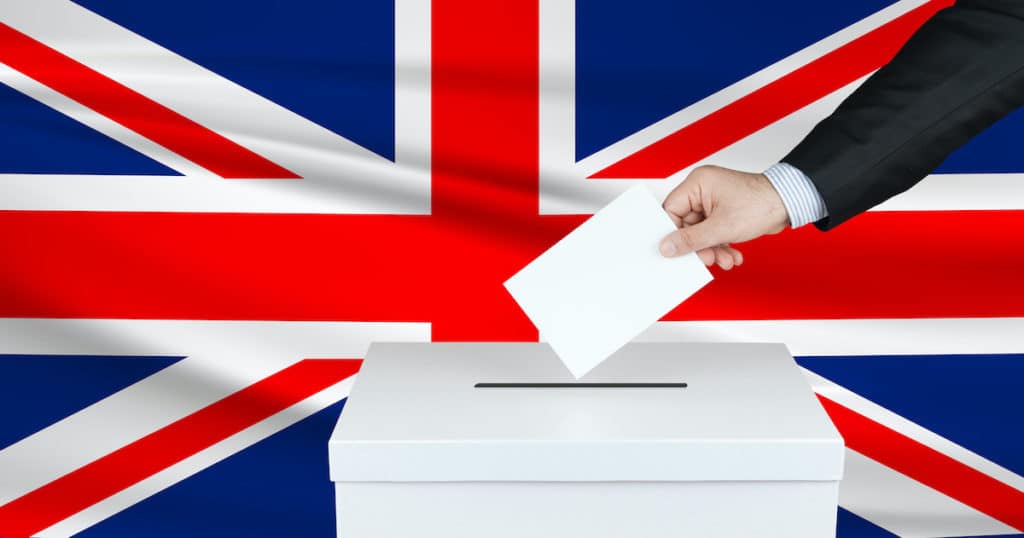 Graphic concept of an election in Great Britain, depicting by the hand of a voter putting his vote in the ballot box with the flag of the United Kingdom in the background. (Photo: AdobeStock)
