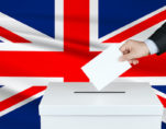 Graphic concept of an election in Great Britain, depicting by the hand of a voter putting his vote in the ballot box with the flag of the United Kingdom in the background. (Photo: AdobeStock)