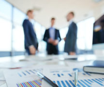 Business statistics and financial reports on an office table with three men talking in the background. (Photo: AdobeStock)