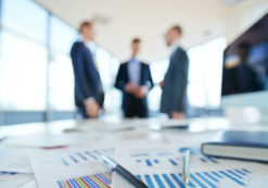 Business statistics and financial reports on an office table with three men talking in the background. (Photo: AdobeStock)