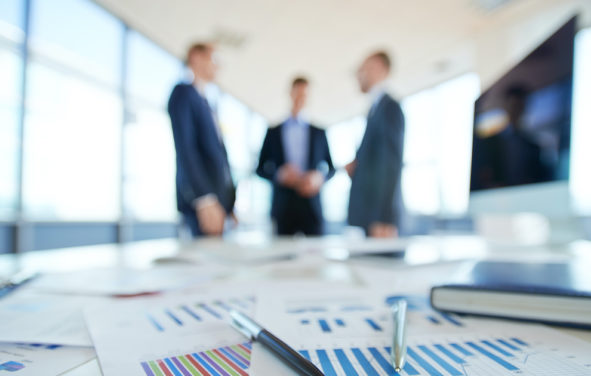 Business statistics and financial reports on an office table with three men talking in the background. (Photo: AdobeStock)