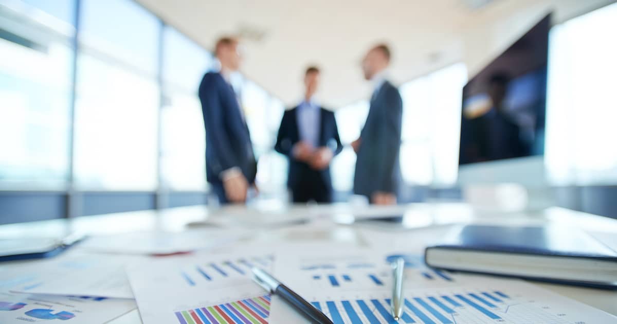 Business statistics and financial reports on an office table with three men talking in the background. (Photo: AdobeStock)