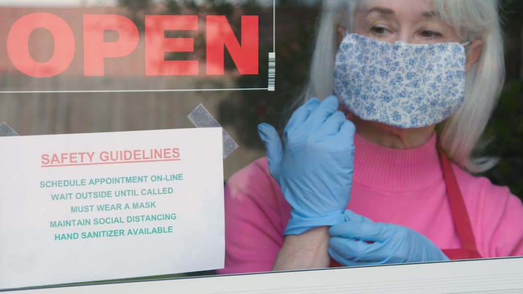 With cautious optimism a small business owner wearing a face mask and disposable gloves posts safety rules as she reopens her store after the coronavirus (COVID-19)shutdown. (Photo: AdobeStock/JHDT Productions)