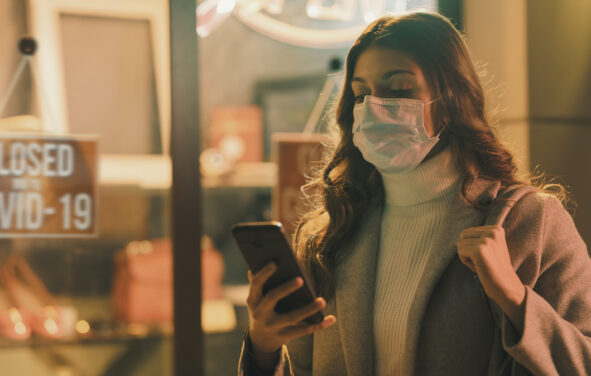 Shop closed due to coronavirus outbreak and woman looking at her mobile device and wearing a surgical mask. (Photo: AdobeStock)
