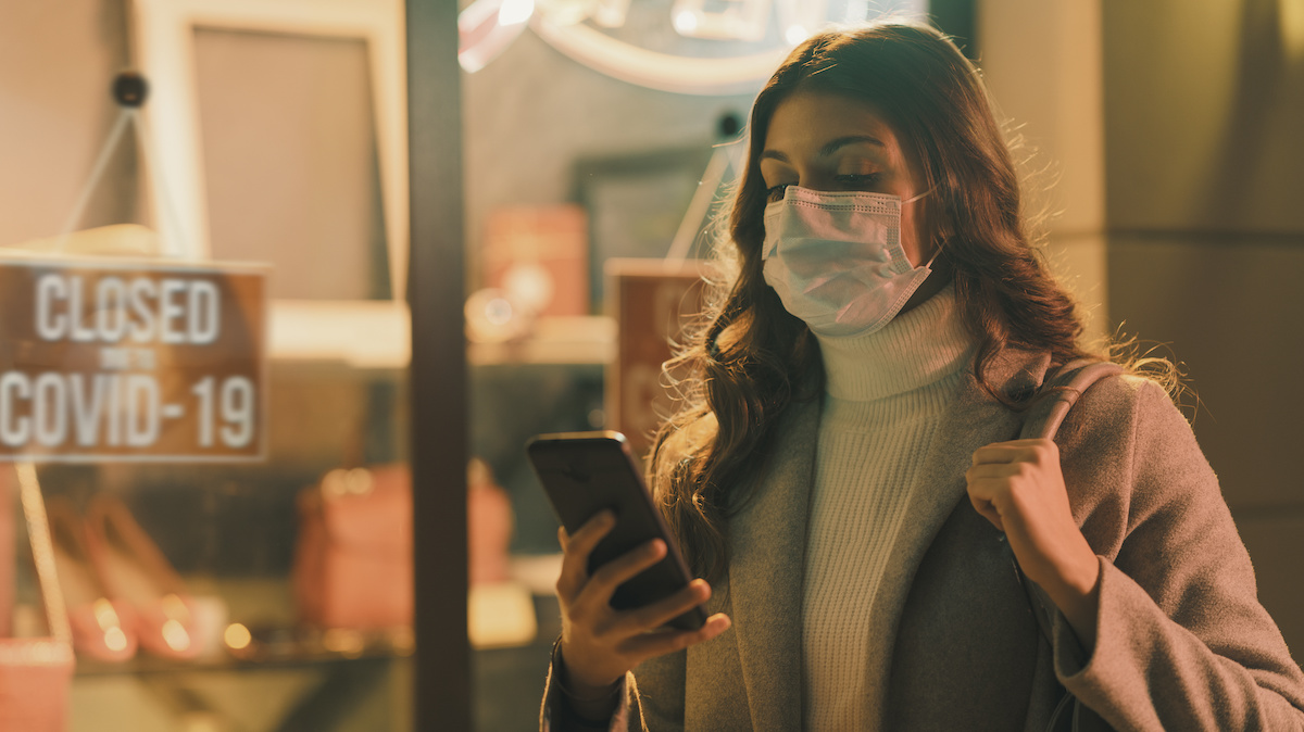 Shop closed due to coronavirus outbreak and woman looking at her mobile device and wearing a surgical mask. (Photo: AdobeStock)